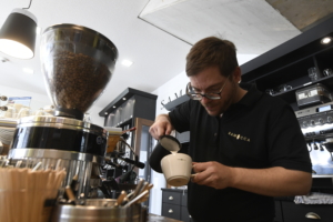 Ein Mann gießt Kaffee um. Er steht in einem Café und benutzt eine Kaffeemaschine.