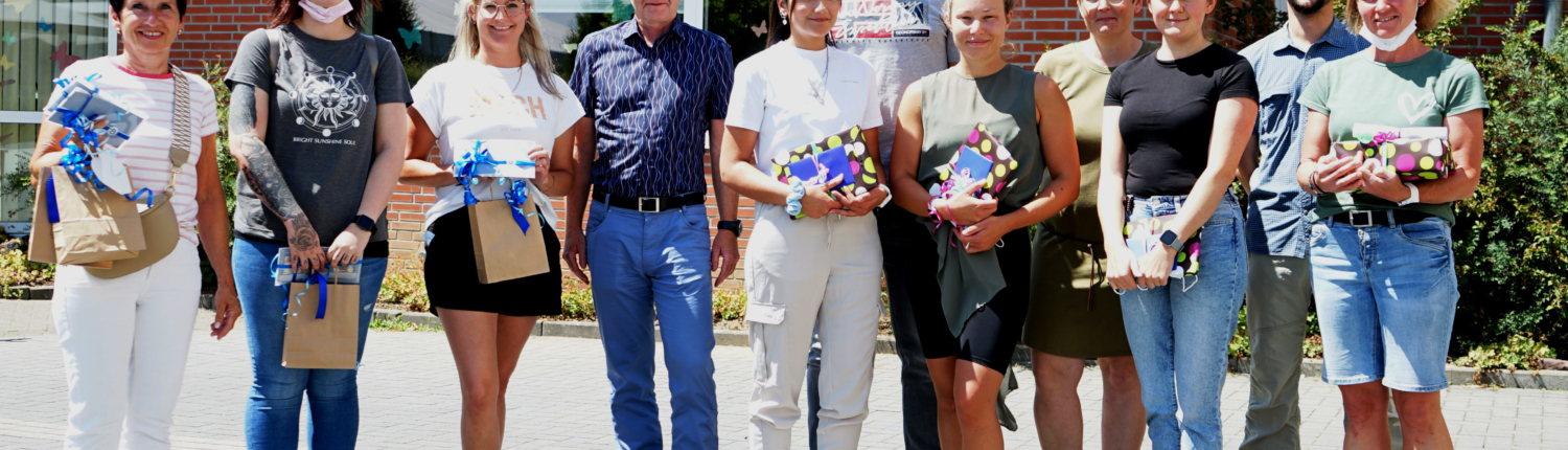 Eine Gruppe von Personen steht draußen vor einem Haus. Sie halten Blumen und kleine Geschenke in der Hand.