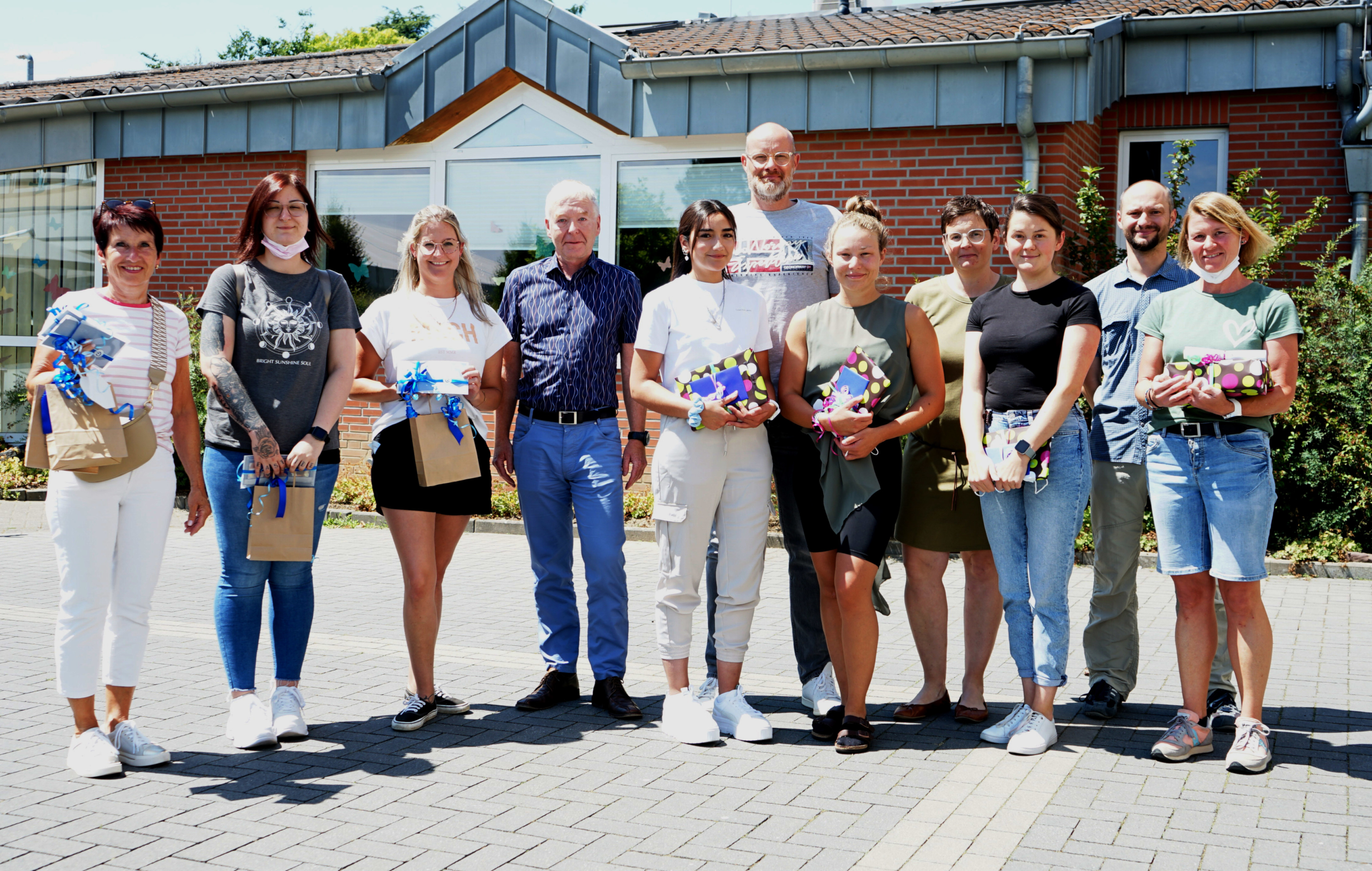 Eine Gruppe von Personen steht draußen vor einem Haus. Sie halten Blumen und kleine Geschenke in der Hand.