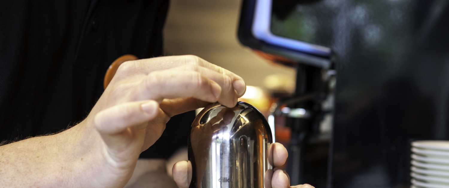 Barista bereitet mit einer Kaffeekanne frisch gemahlenen Kaffee in einem modernen Café zu, während ein Computer im Hintergrund sichtbar ist.