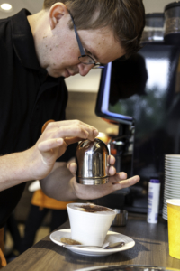 Barista bereitet mit einer Kaffeekanne frisch gemahlenen Kaffee in einem modernen Café zu, während ein Computer im Hintergrund sichtbar ist.