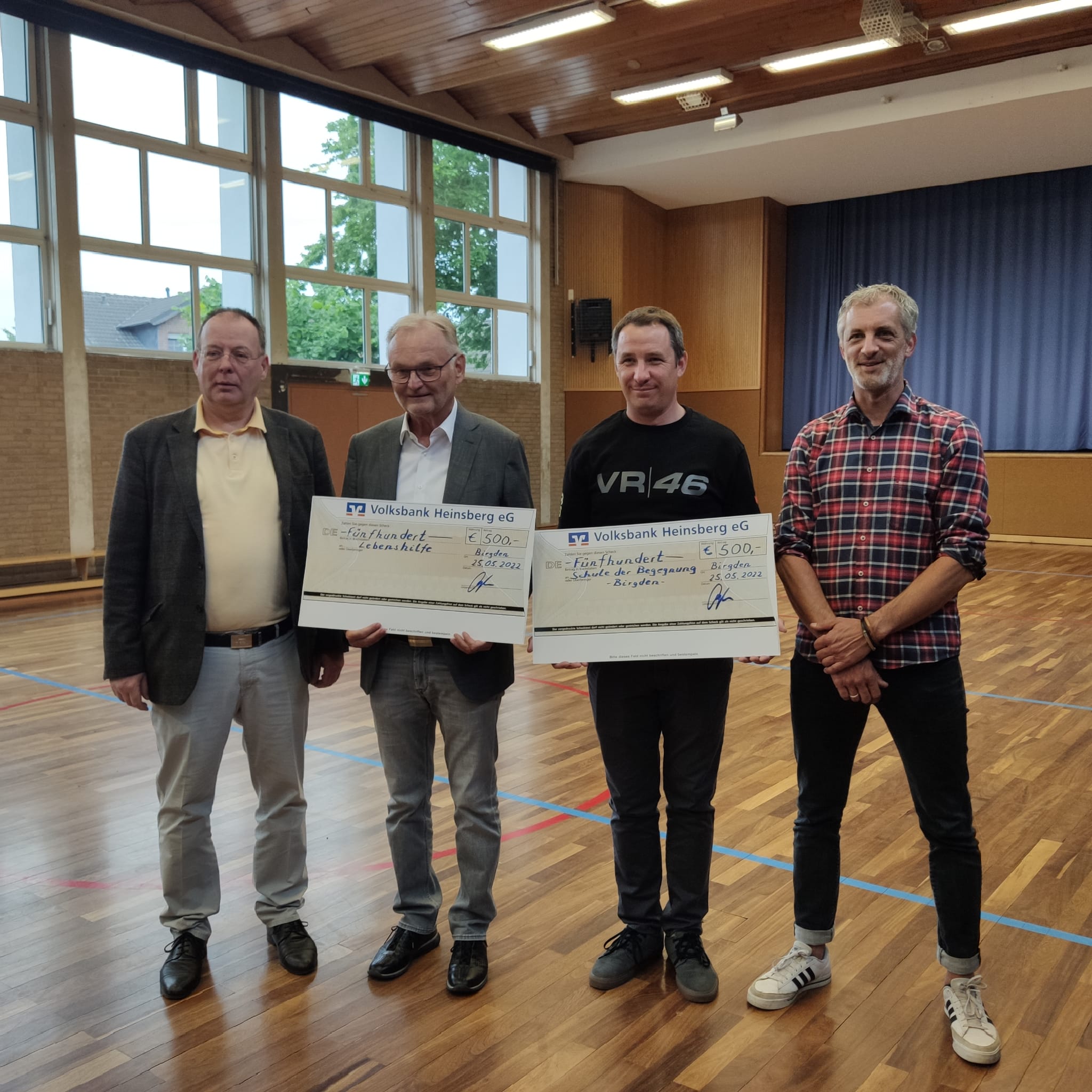 Vier Personen stehen in einem Saal und halten Urkunden oder Auszeichnungen in den Händen. Der Raum hat große Fenster und Holzboden. Die Veranstaltung scheint eine Ehrung oder Preisverleihung zu sein.