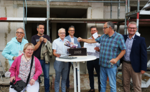 Gruppenfoto von Bauarbeitern und Unterstützern bei einer feierlichen Veranstaltung, die den Fortschritt eines Bauprojekts symbolisiert. Im Vordergrund steht ein Tisch mit einer Blumenschale, während die Teilnehmer lächeln und an einem Bauort posieren.