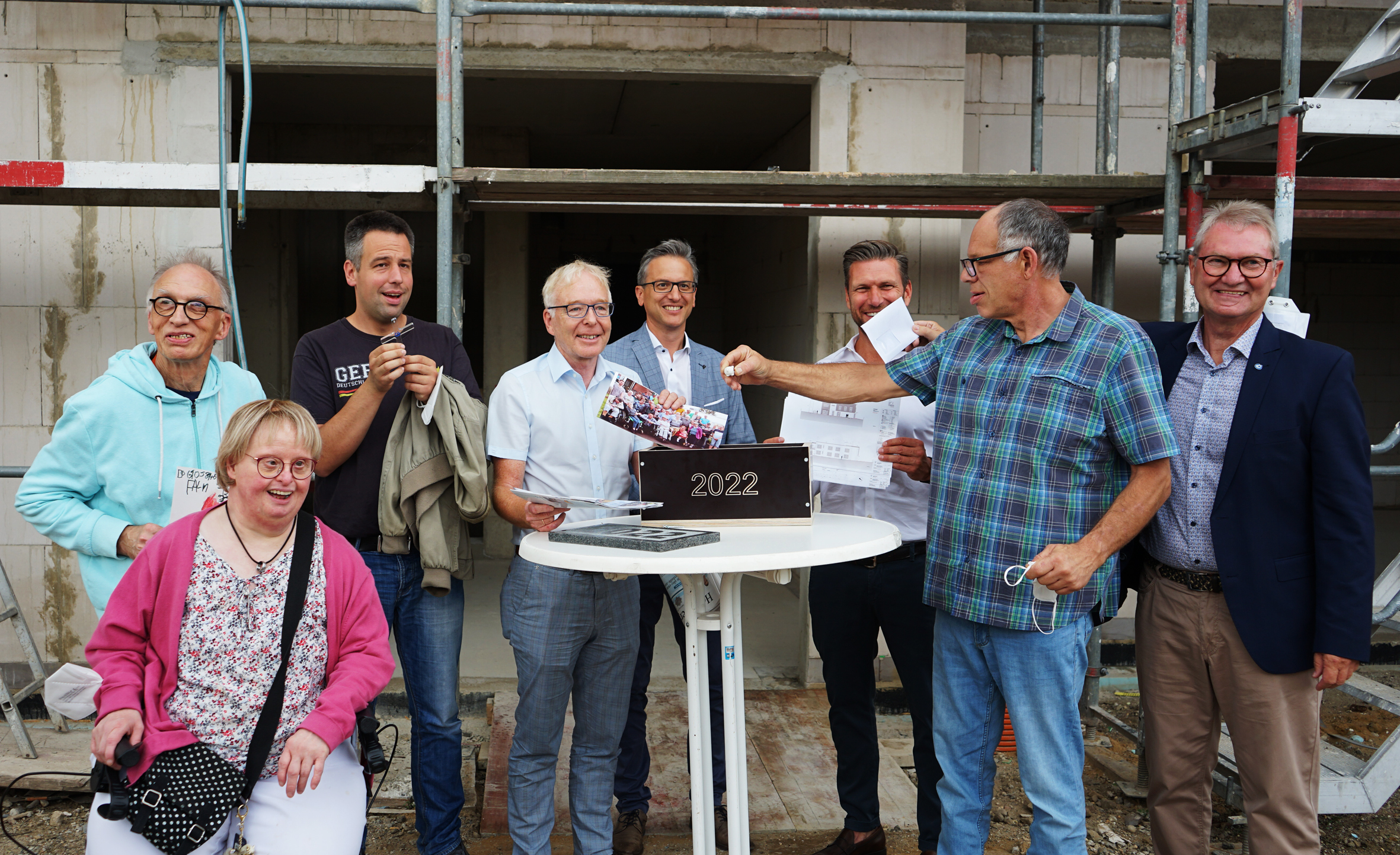 Gruppenfoto von Bauarbeitern und Unterstützern bei einer feierlichen Veranstaltung, die den Fortschritt eines Bauprojekts symbolisiert. Im Vordergrund steht ein Tisch mit einer Blumenschale, während die Teilnehmer lächeln und an einem Bauort posieren.