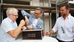 Drei Männer arbeiten gemeinsam an einer Holzkiste mit der Aufschrift "2022", während sie Werkzeuge verwenden. Im Hintergrund sind Baugerüste sichtbar, was auf eine Renovierung oder Bauarbeiten hindeutet.