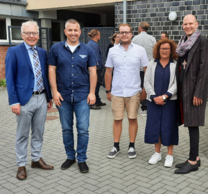 Gruppenfoto von fünf Personen vor einem modernen Gebäude, die in informeller Kleidung lächeln und sich für die Kamera aufstellen.