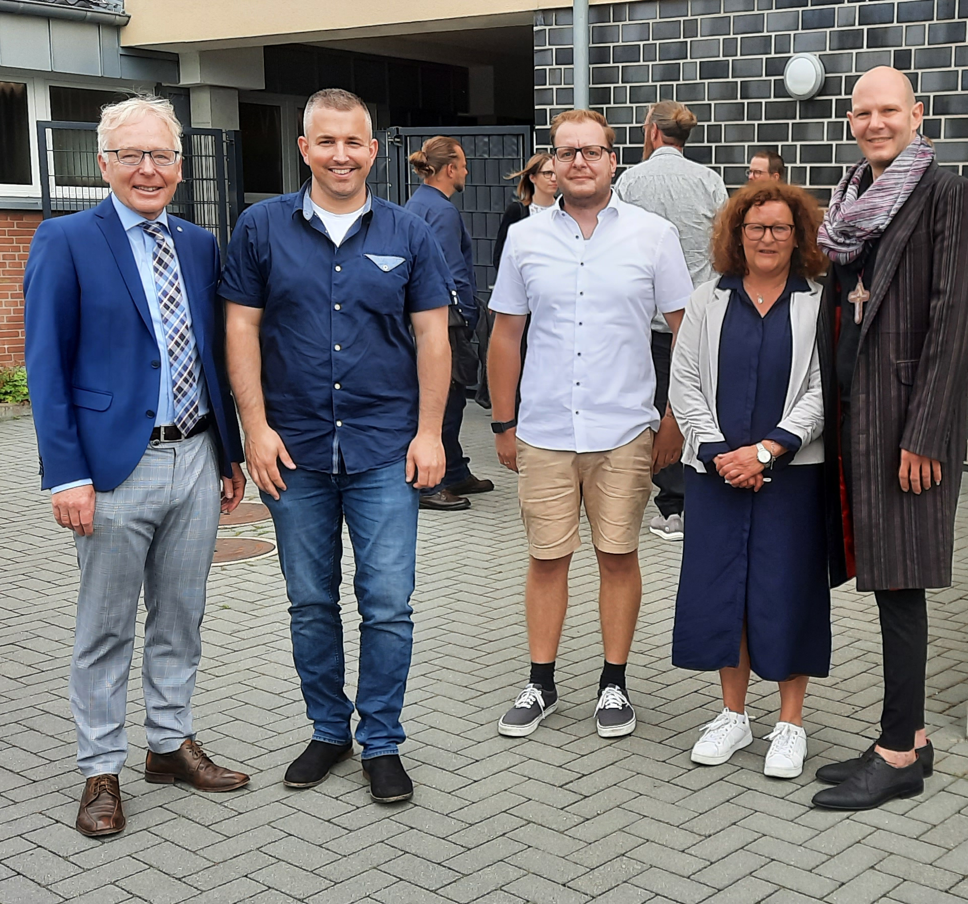 Gruppenfoto von fünf Personen vor einem modernen Gebäude, die in informeller Kleidung lächeln und sich für die Kamera aufstellen.