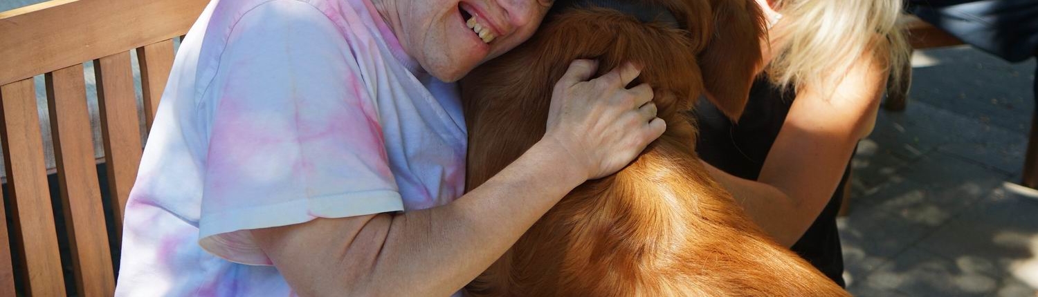Ein Mann umarmt einen Hund. Der Hund ist rotbraun. Beide lächeln.