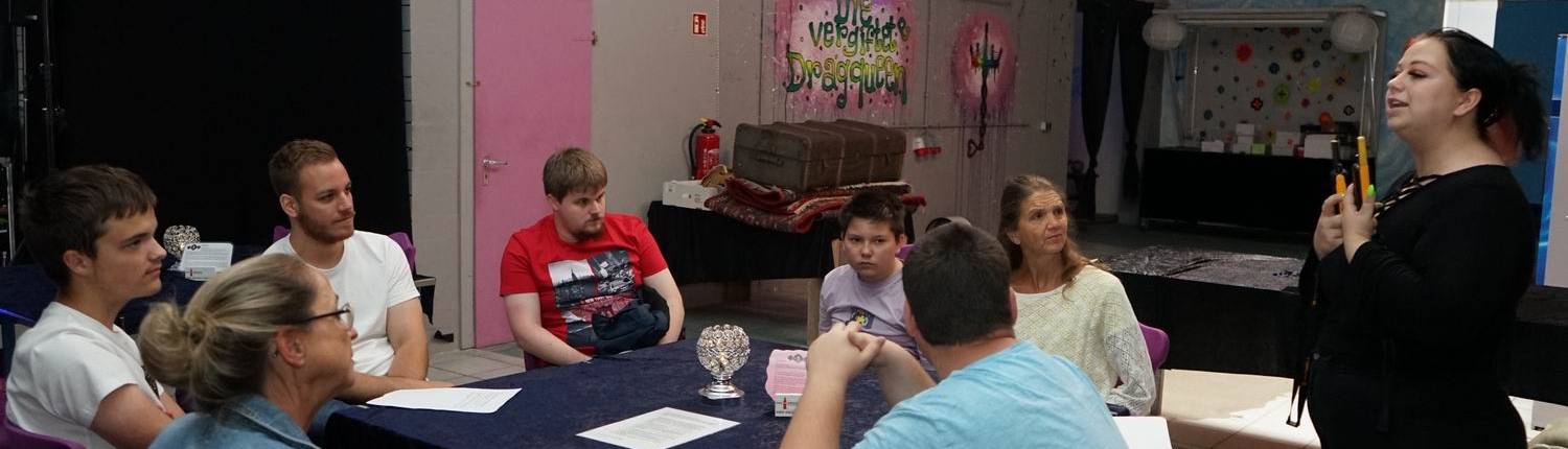 Eine Gruppe von Personen sitzt an einem Tisch in einem Veranstaltungsraum. Im Hintergrund sind farbige Wände und Dekorationen sichtbar. Die Teilnehmer scheinen an einer Diskussion oder einem Meeting teilzunehmen.