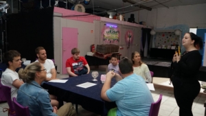 Eine Gruppe von Personen sitzt an einem Tisch in einem Veranstaltungsraum. Im Hintergrund sind farbige Wände und Dekorationen sichtbar. Die Teilnehmer scheinen an einer Diskussion oder einem Meeting teilzunehmen.