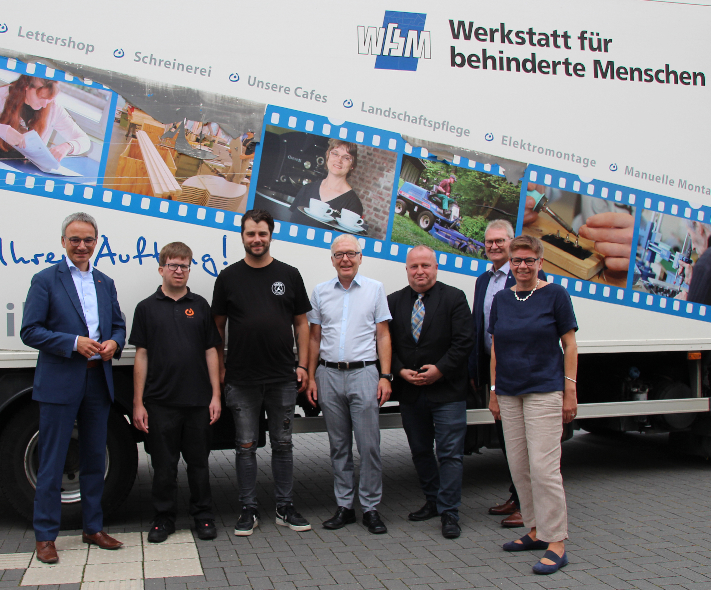 Gruppenfoto von Mitarbeitern des Werkstatt für behinderte Menschen vor einem Fahrzeug, das mit Informationen und Bildern zur Einrichtung gestaltet ist.
