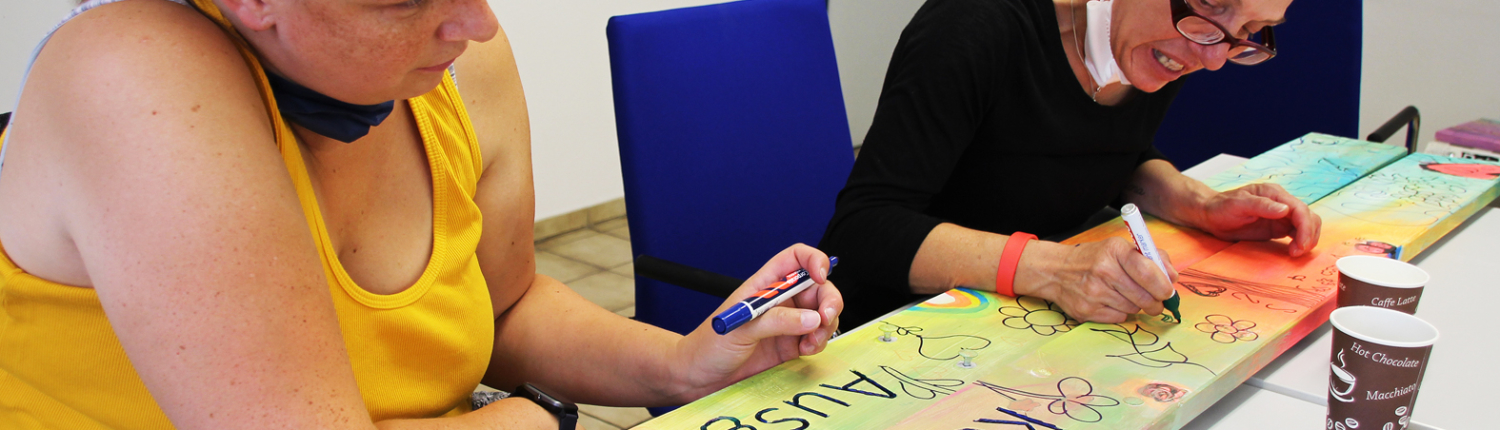 Zwei Frauen sitzen an einem Tisch und gestalten ein farbenfrohes Banner mit verschiedenen Schriftzügen. Die Frau links trägt ein gelbes Oberteil und die Frau rechts ein schwarzes Oberteil. Beide konzentrieren sich auf ihre Malerei.