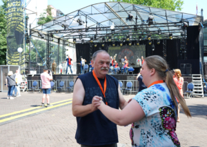 Zwei Personen tanzen und feiern auf einem Festivalgelände vor einer Bühne, umgeben von anderen Besuchern.