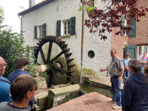 Eine Wassermühle mit einem großen Rad. Ein Mann zeigt etwas. Es stehen Leute davor.