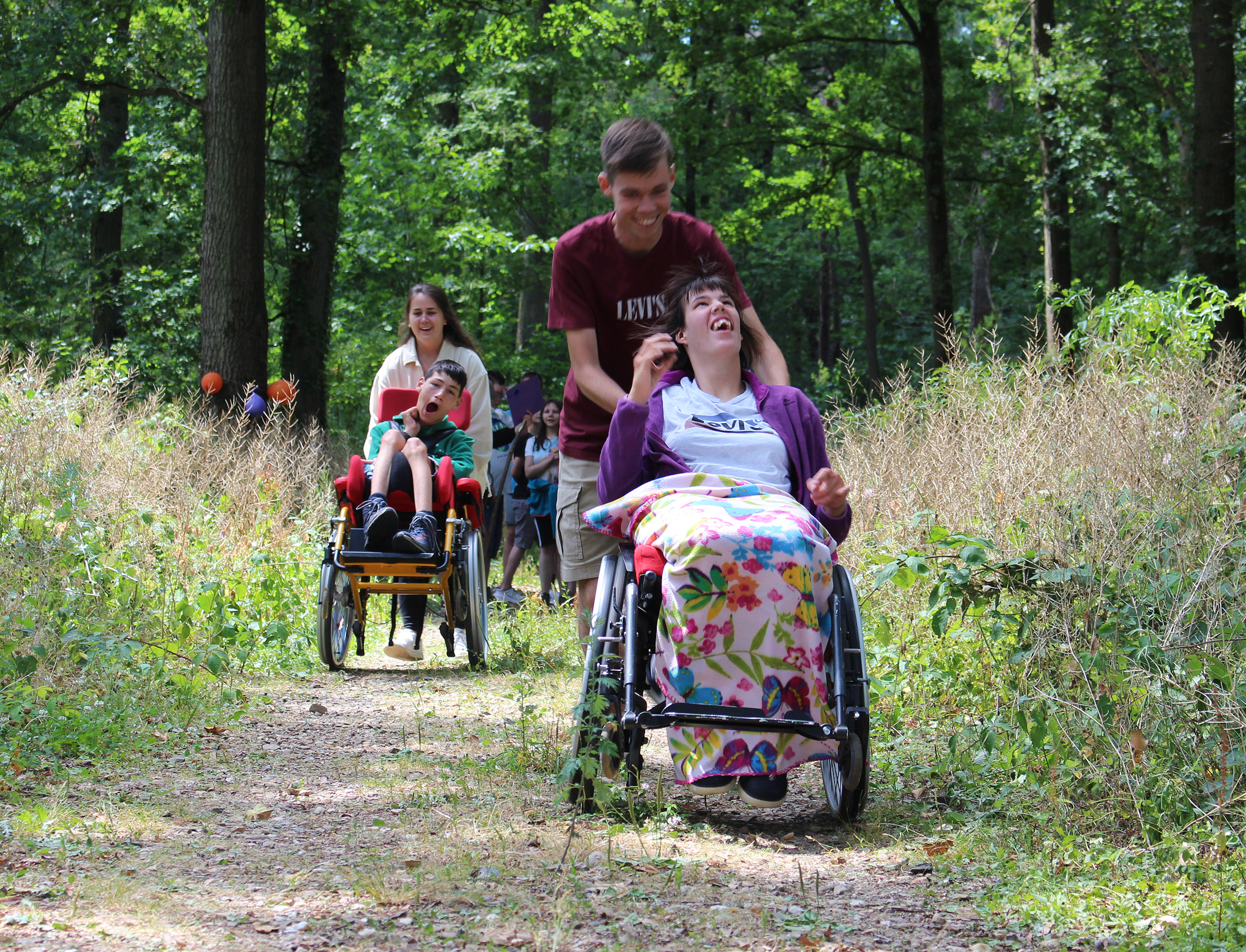 Gruppen von Menschen im Rollstuhl, die einen schmalen Waldweg entlangfahren, während sie von Freunden oder Betreuern geschoben werden. Die Szene zeigt eine inklusive Outdoor-Aktivität in der Natur.