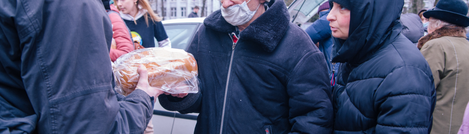 Eine ältere Person erhält Lebensmittelhilfe von Freiwilligen in einer urbanen Umgebung, während andere Menschen im Hintergrund stehen.