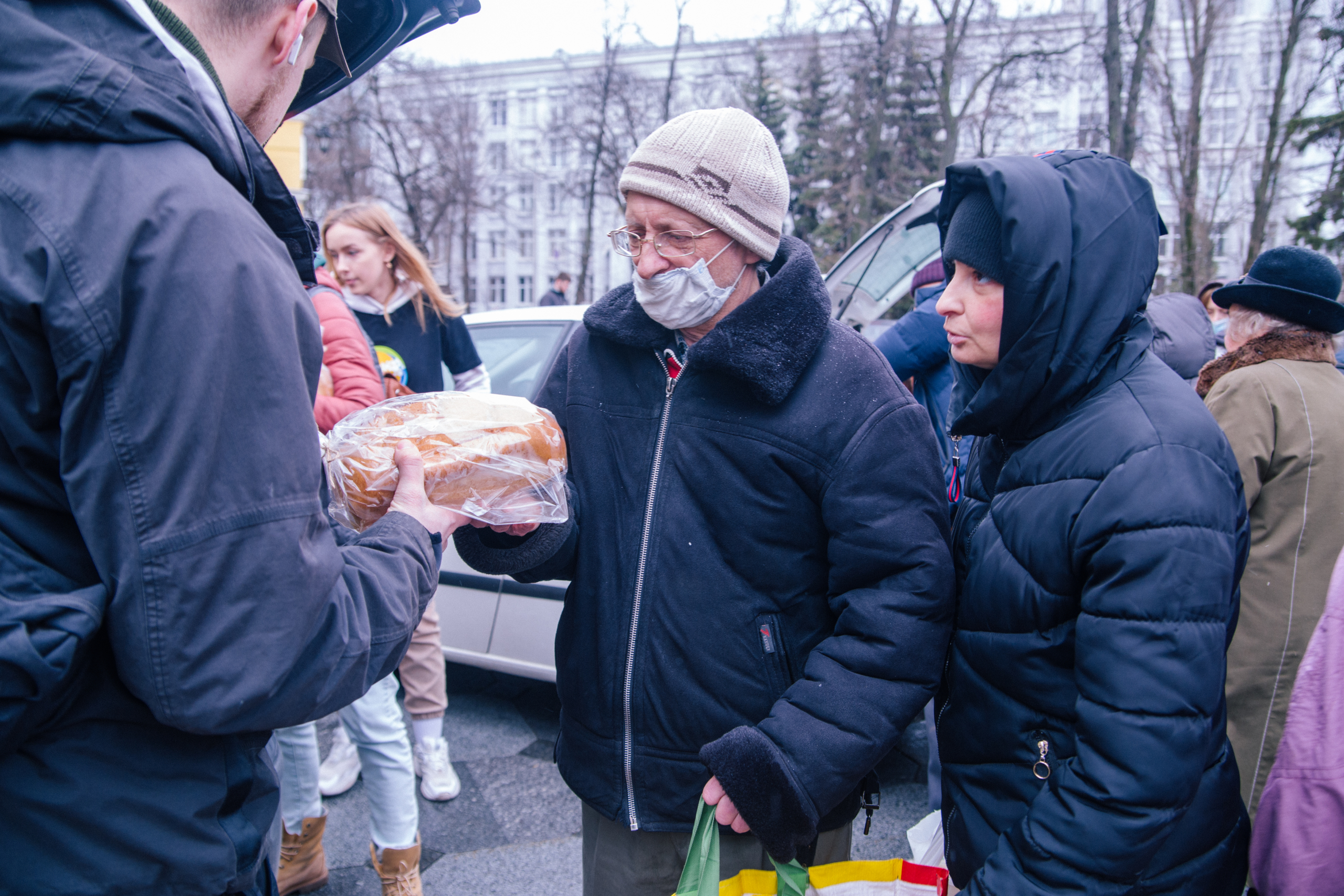 Eine ältere Person erhält Lebensmittelhilfe von Freiwilligen in einer urbanen Umgebung, während andere Menschen im Hintergrund stehen.