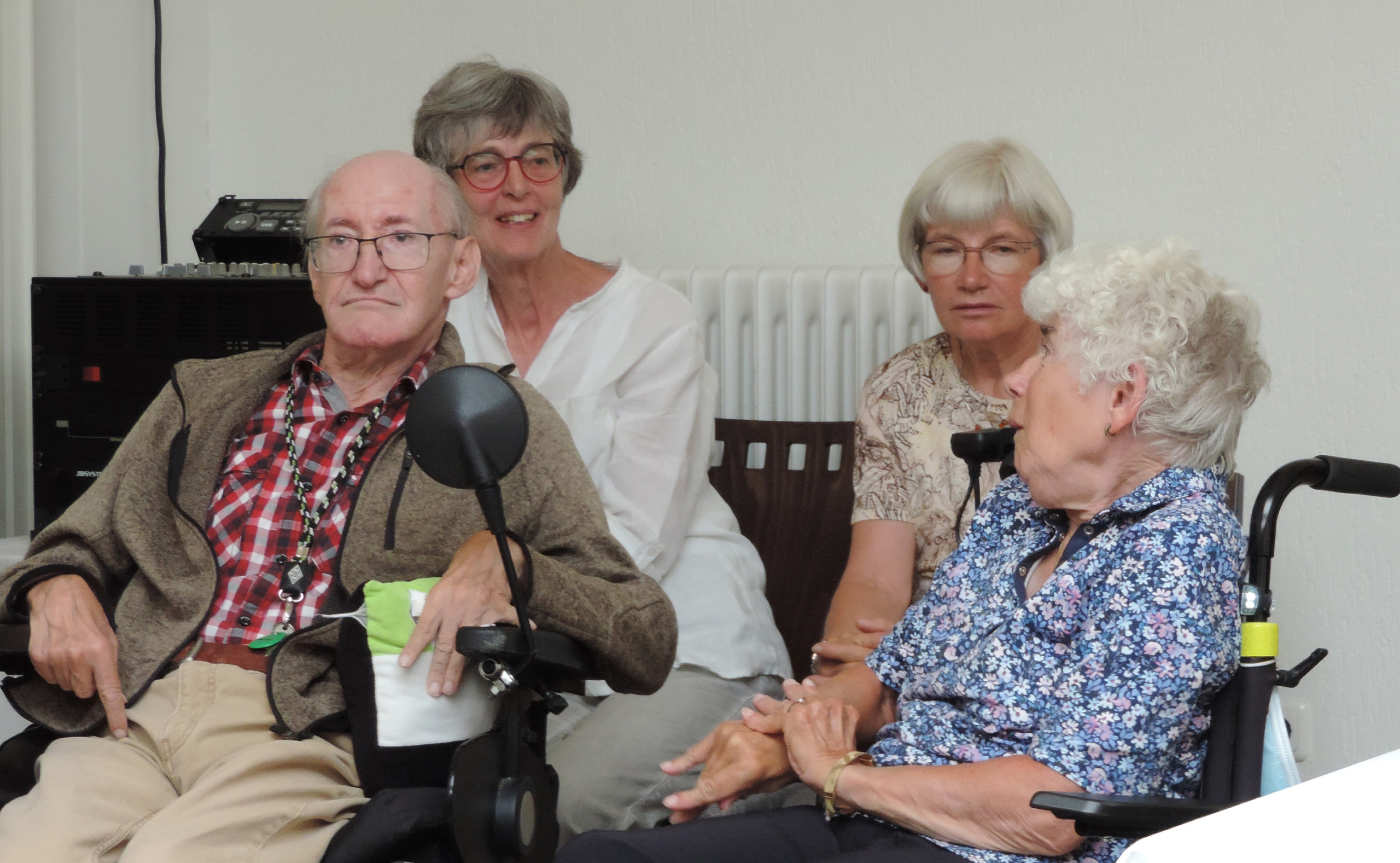 Ein Mann sitzt im Rollstuhl. Zwei Frauen sitzen daneben. Sie sind in einem Raum mit einem Tisch und einem Regal.
