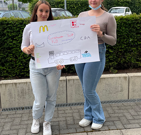 Zwei Jugendliche halten ein großes Plakat, auf dem Informationen oder Botschaften zu sehen sind, vor einer grünen Hecke. Sie tragen legere Kleidung und stehen auf einem gepflasterten Weg.