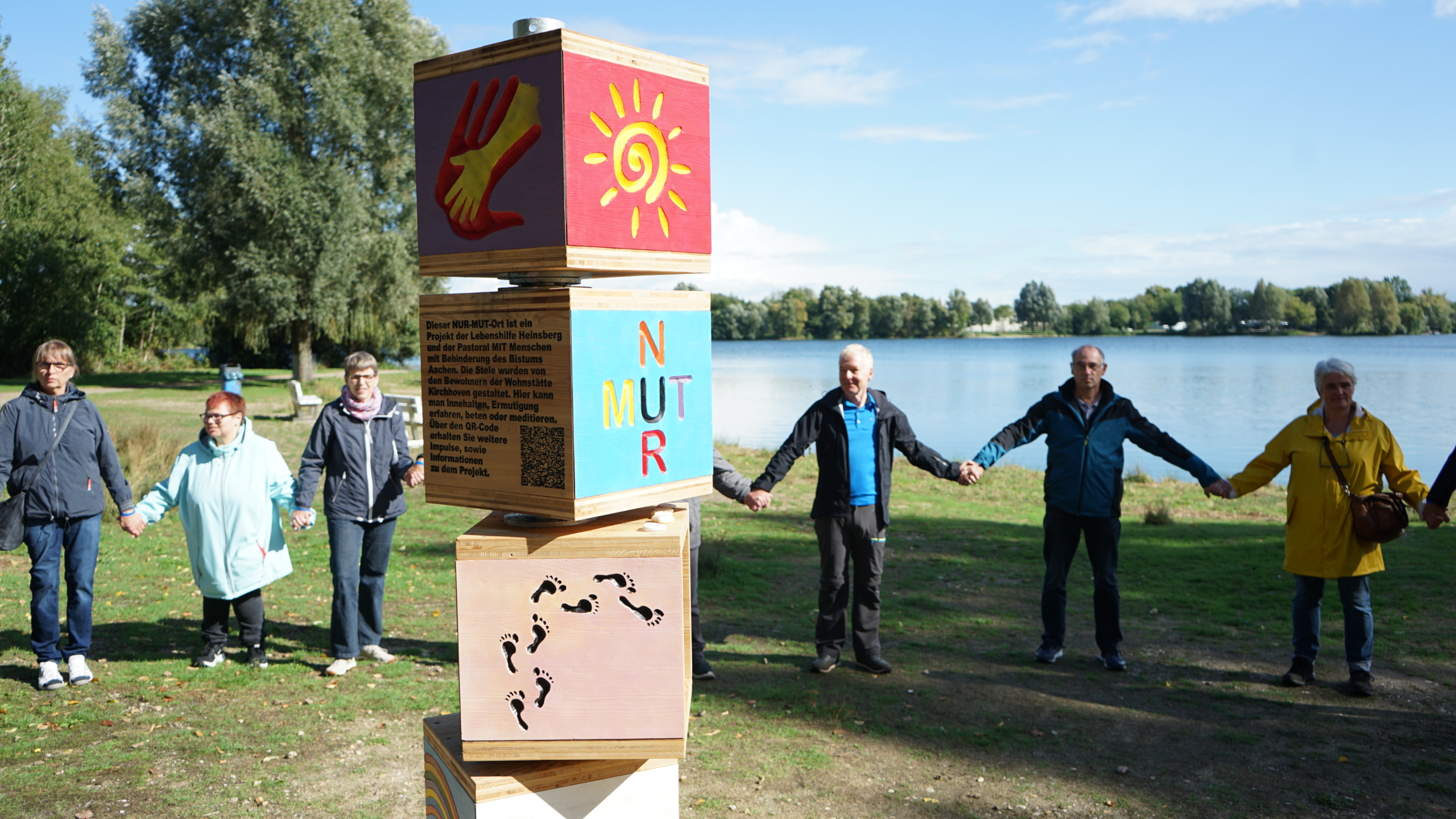 Menschen halten Händchen. Sie stehen im Freien vor einem See.