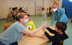 Zwei Kinder geben sich die Fäuste. Eines hat ein grünes T-Shirt, das andere ein schwarzes. Sie stehen an einem Tisch.