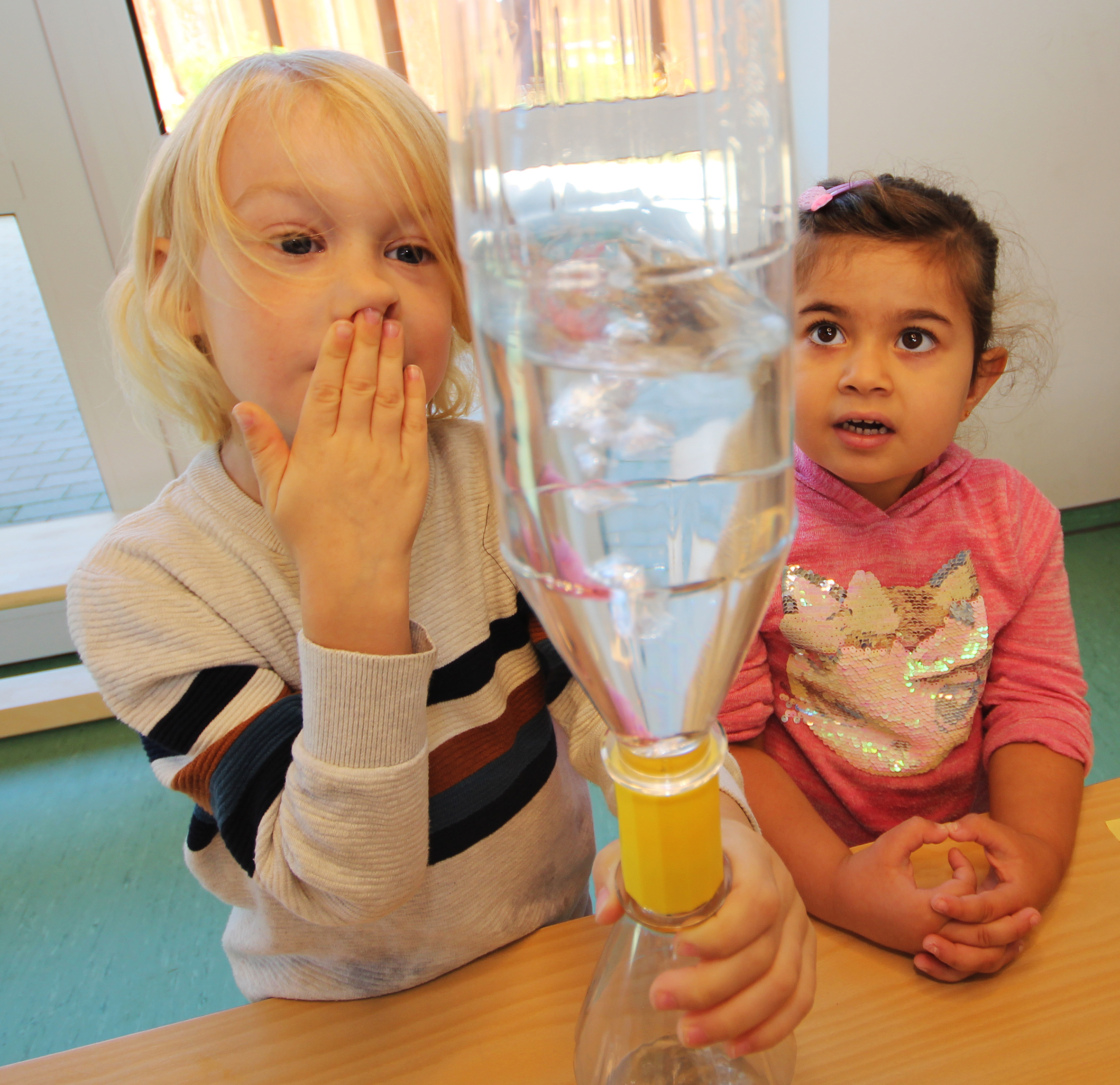 Zwei Kinder sitzen an einem Tisch und beobachten ein Experiment mit einer transparenten Flasche, die Wasser enthält. Das blonde Kind hält die Flasche und schaut neugierig, während das dunkelhaarige Kind daneben sitzt und interessiert zusieht.