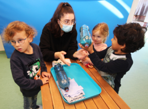 Eine Betreuerin zeigt Kindern in einem Lernumfeld eine Aktivität mit einem blauen Spielzeugboot. Die Kinder sind neugierig und interagieren miteinander, während sie die Materialien auf einem Tisch erkunden.