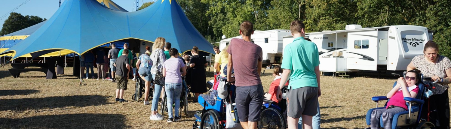 Menschen stehen in einer Schlange vor einem blauen Zelt auf einem Festivalgelände, umgeben von Wohnwagen und Bäumen.
