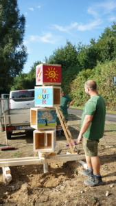 Ein Mann steht auf einer Baustelle und stapelt bemalte Holzboxen mit verschiedenen Motiven. Im Hintergrund ist ein Fahrzeug und Bäume zu sehen.