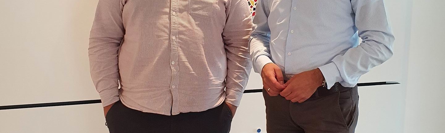 Zwei Männer stehen nebeneinander. Einer trägt ein weißes Hemd, der andere ein blaues. Beide stehen vor einer Wand mit bunten Punkten.