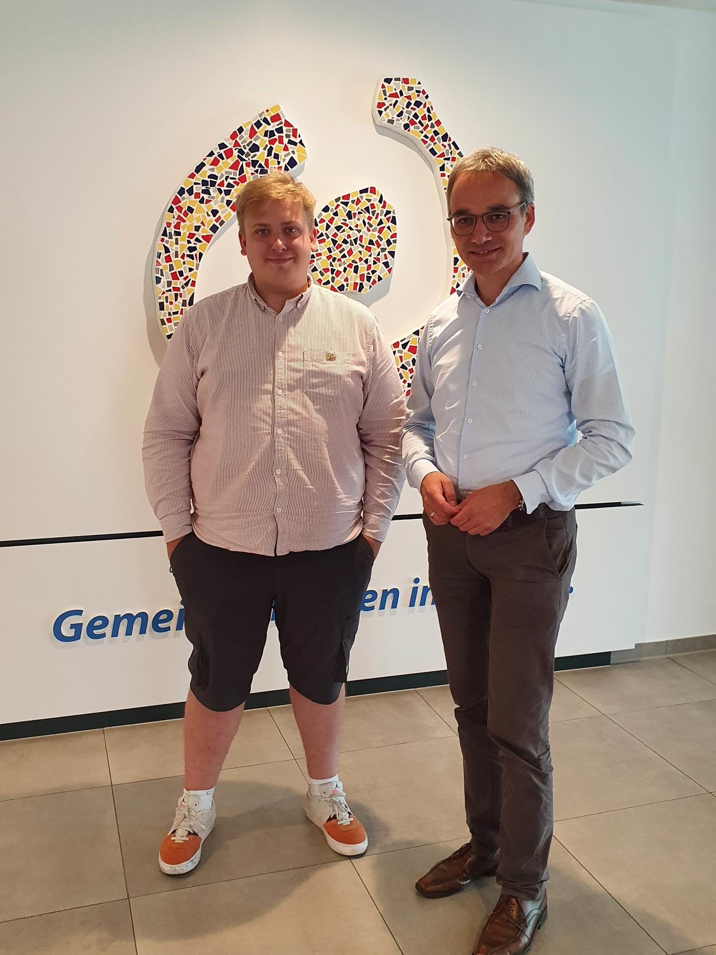 Zwei Männer stehen nebeneinander. Einer trägt ein weißes Hemd, der andere ein blaues. Beide stehen vor einer Wand mit bunten Punkten.