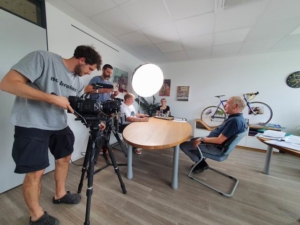 Ein Mann filmt mit einer Kamera. Er steht vor einer Gruppe von Menschen. Es gibt viele Fahrräder im Hintergrund.