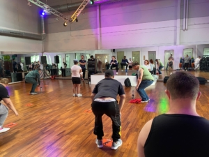 Menschen machen Sport in einem hellen Raum. Sie tanzen oder machen Übungen. Es gibt große Spiegel an der Wand.
