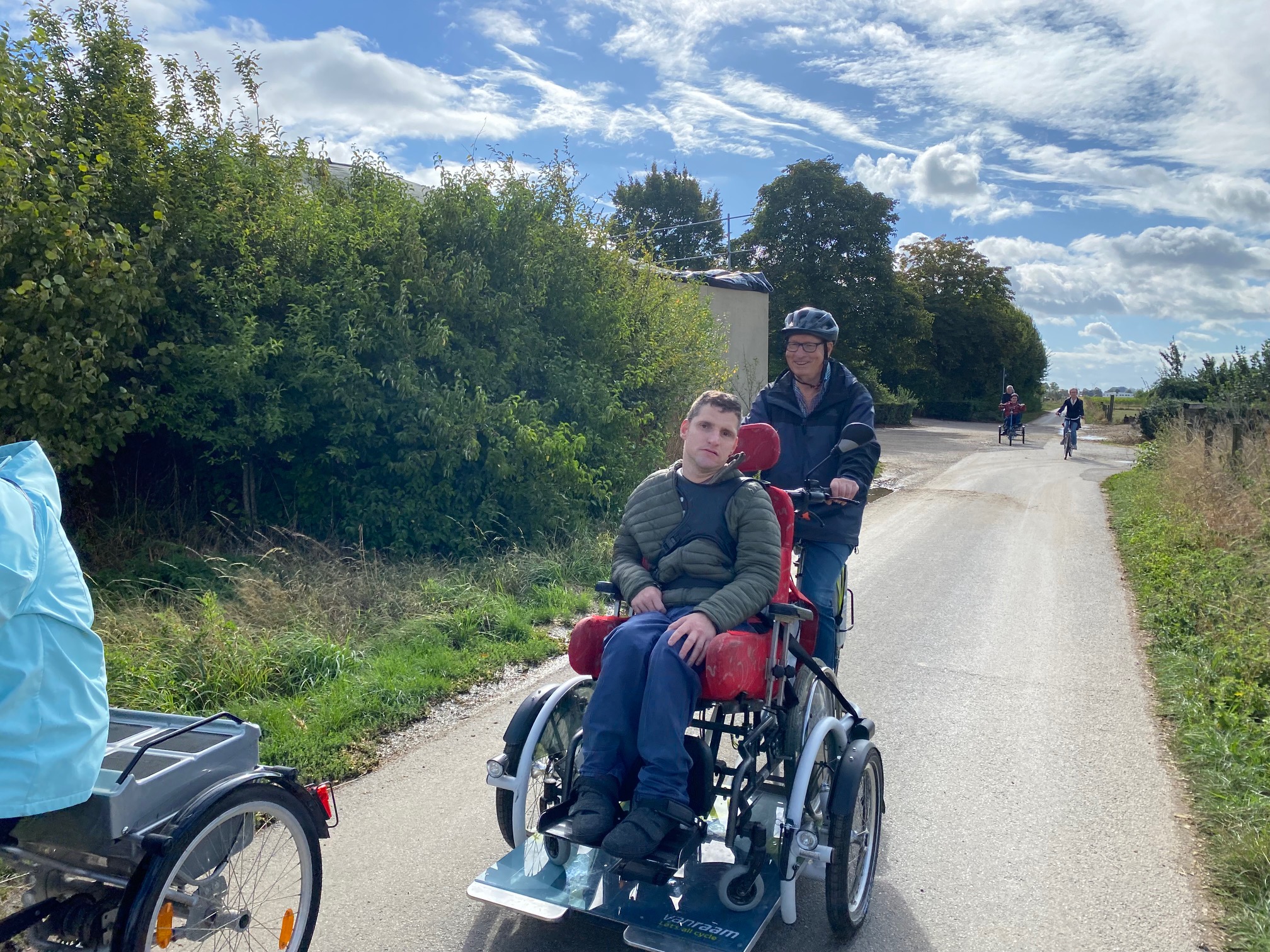Zwei Personen fahren auf einem modernen Dreirad auf einem Radweg, umgeben von grüner Landschaft und unter blauem Himmel.