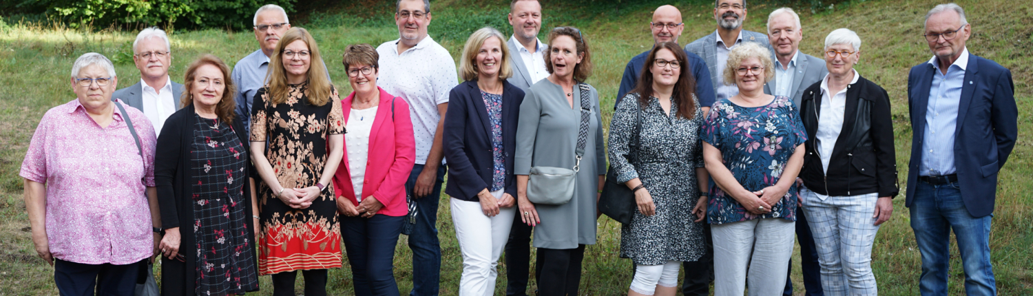 Gruppenfoto von 16 Personen im Freien, umgeben von Grünflächen und Bäumen, die lächeln und in lässiger Kleidung posieren. Ideal für Inhalte über Teamarbeit, Gemeinschaft oder Veranstaltungen.