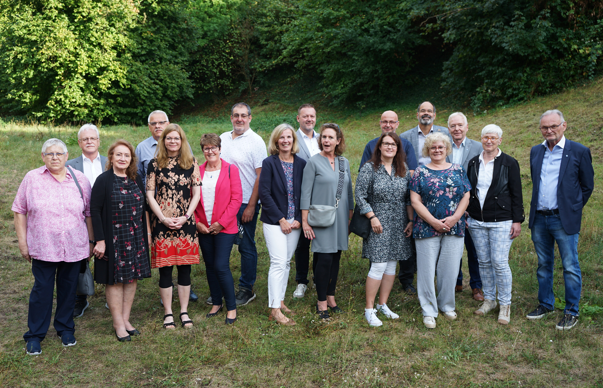 Gruppenfoto von 16 Personen im Freien, umgeben von Grünflächen und Bäumen, die lächeln und in lässiger Kleidung posieren. Ideal für Inhalte über Teamarbeit, Gemeinschaft oder Veranstaltungen.