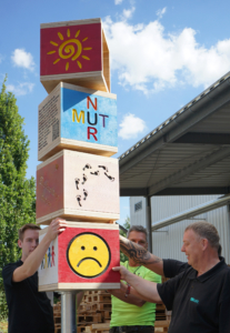 Drei Männer bauen einen hohen Turm aus Holzbrettern. Sie stehen draußen vor einem Gebäude.