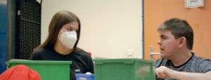 Eine Person mit Mundschutz arbeitet an einem Tisch mit farbigen Behältern in einem Raum, der für organisatorische oder kreative Tätigkeiten genutzt wird.