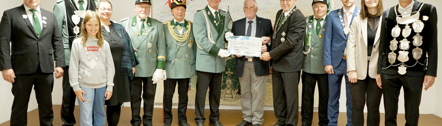 Gruppenfoto einer Zeremonie mit sieben festlich gekleideten Personen in traditionellen Uniformen, die eine Urkunde überreichen. Im Hintergrund eine neutrale Wand, die Veranstaltung findet in einem Innenraum statt.