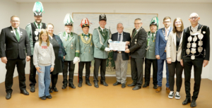 Gruppenfoto einer Zeremonie mit sieben festlich gekleideten Personen in traditionellen Uniformen, die eine Urkunde überreichen. Im Hintergrund eine neutrale Wand, die Veranstaltung findet in einem Innenraum statt.