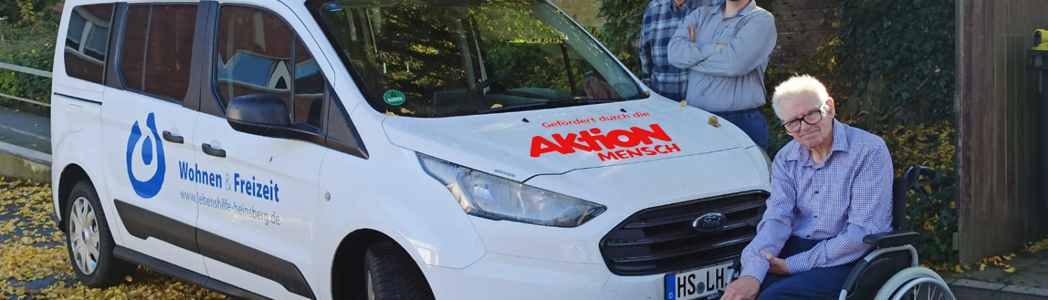 Zwei Männer stehen vor einem weißen Auto. Ein Mann sitzt im Rollstuhl.