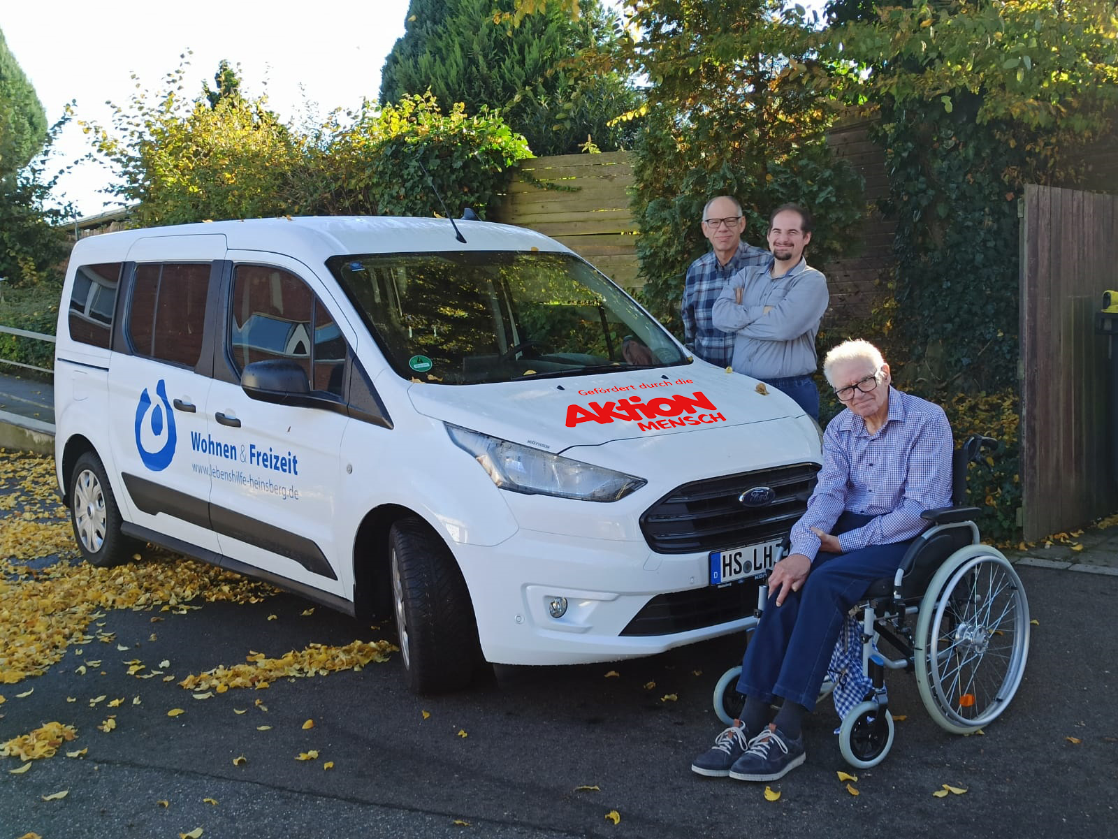 Zwei Männer stehen vor einem weißen Auto. Ein Mann sitzt im Rollstuhl.