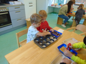 Zwei Kinder spielen in einem hellen Raum mit Spielzeug auf einem Tisch und nutzen eine Backform. Im Hintergrund sind weitere Spielzeuge und eine Erzieherin zu sehen.