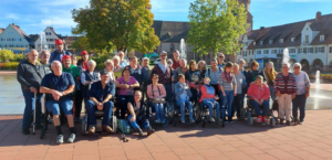 Eine Gruppe von Menschen steht vor einem Brunnen. Es sind viele ältere Menschen dabei.