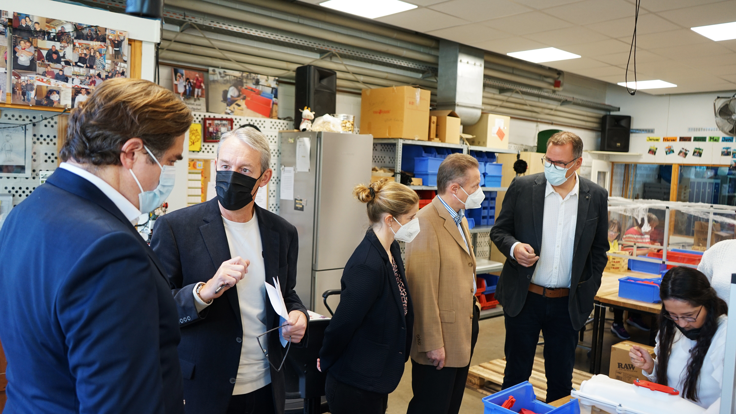 Personen in einem modernen Geschäftsumfeld, die miteinander sprechen und Masken tragen. Im Hintergrund sind Regale mit verschiedenen Produkten zu sehen, was auf eine geschäftliche Besprechung oder Veranstaltung hinweist.