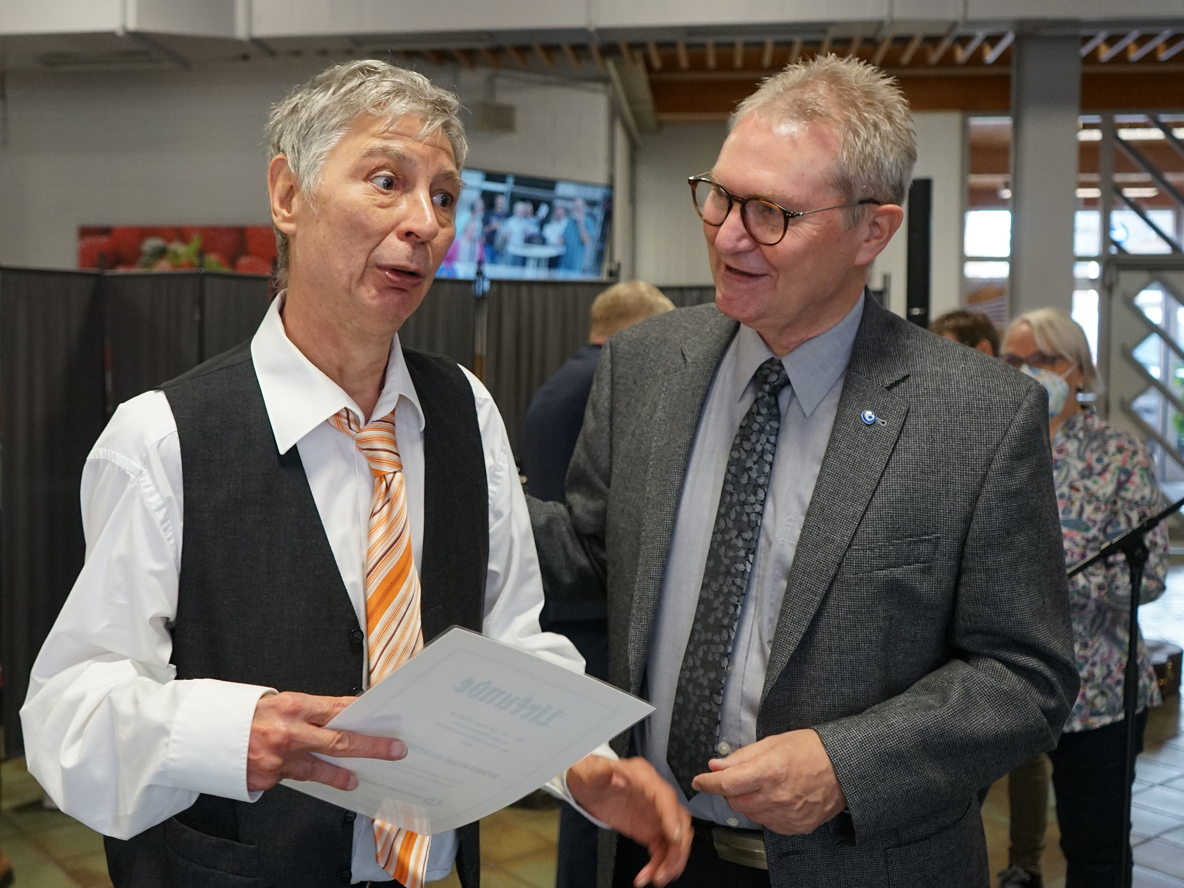 Zwei Männer sprechen miteinander. Einer hält ein Blatt Papier. Sie tragen Anzüge und Krawatten.