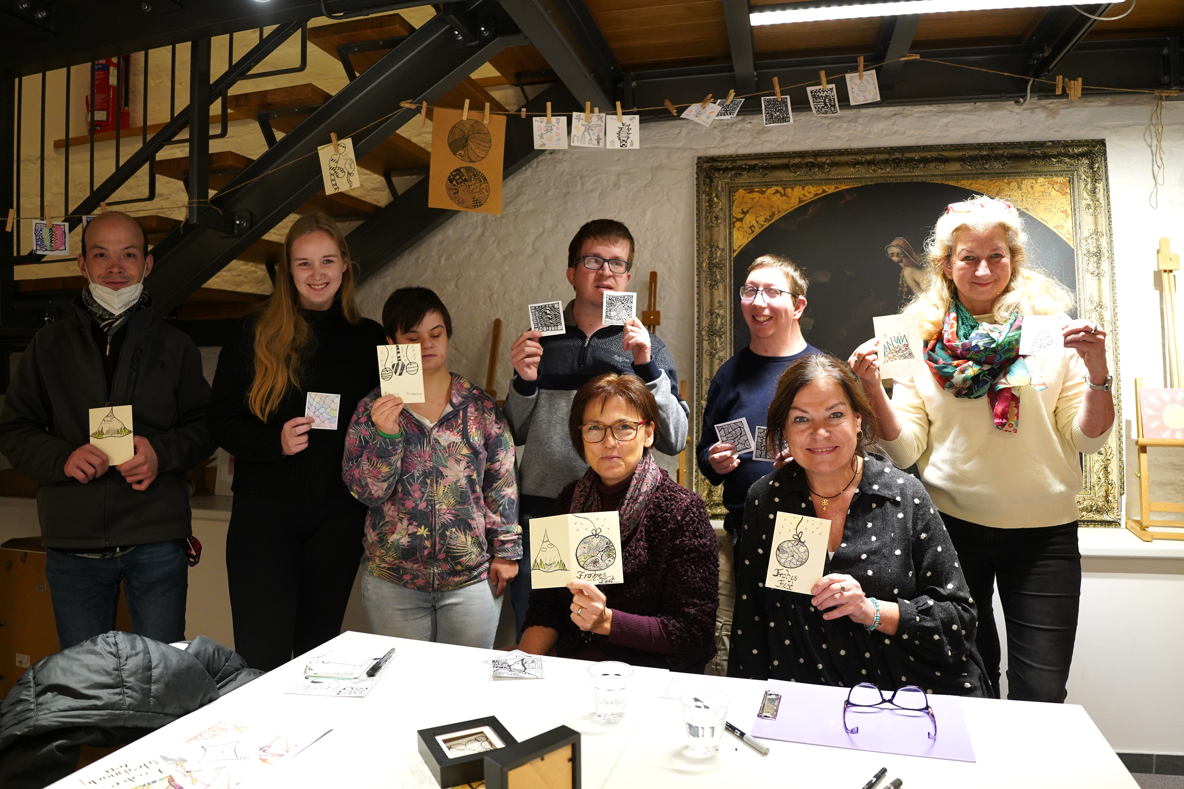 Gruppe von sieben Personen, die in einem gemütlichen Raum zusammenkommen, während sie stolz ihre Kunstwerke oder Bastelprojekte präsentieren. Der Tisch ist mit Materialien und Werkzeugen zum kreativen Arbeiten gedeckt.