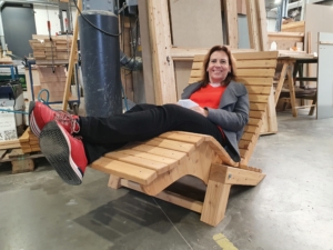 Frau sitzt entspannt auf einem modernen Holzliegestuhl in einem Möbelgeschäft, umgeben von Holzwerkstoffen und Möbeln.