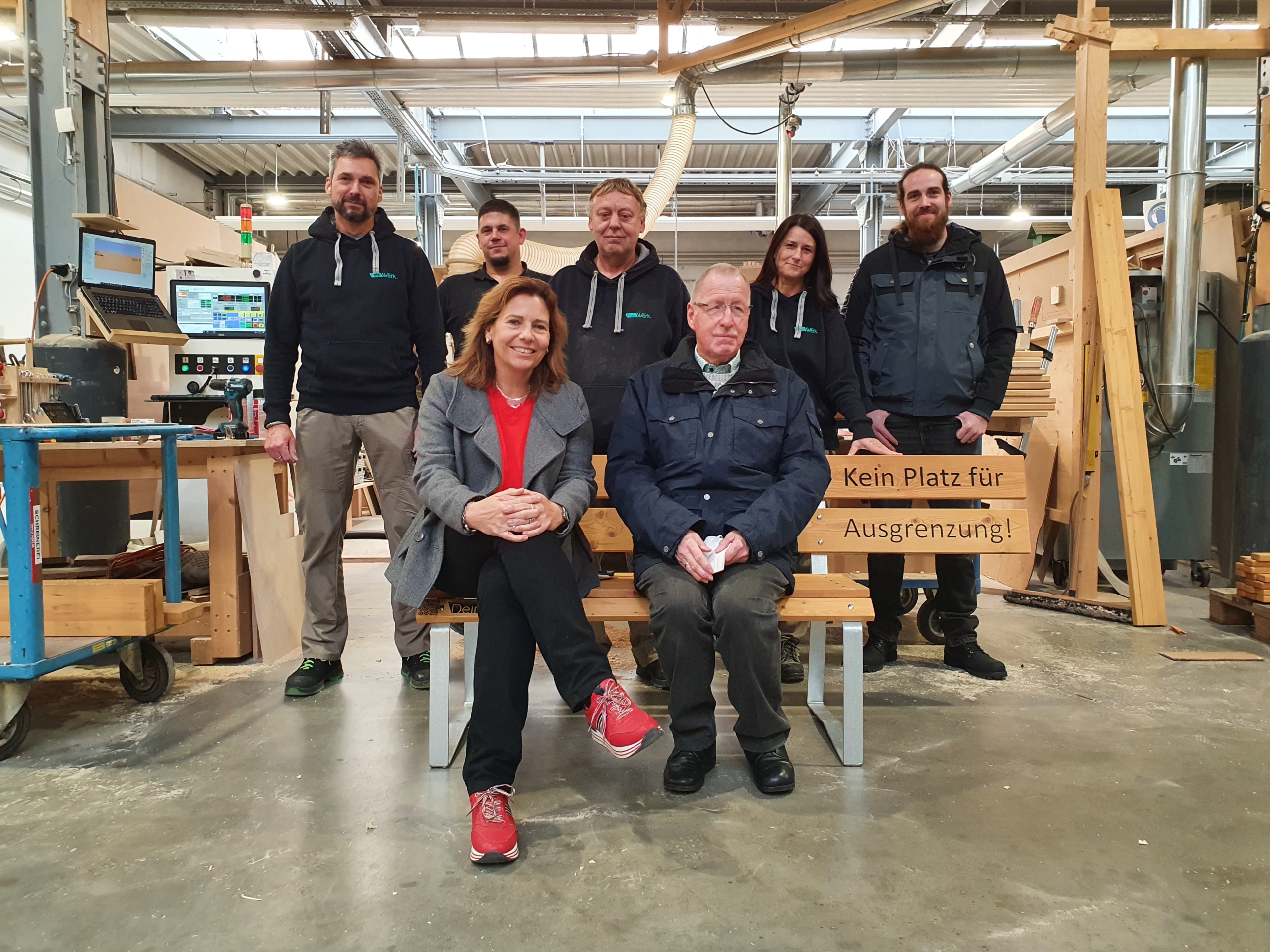 Gruppe von sieben Personen in einer Werkstatt, die auf Holzbänken sitzen und stehen, umgeben von Holzmaterialien und Werkzeugen.