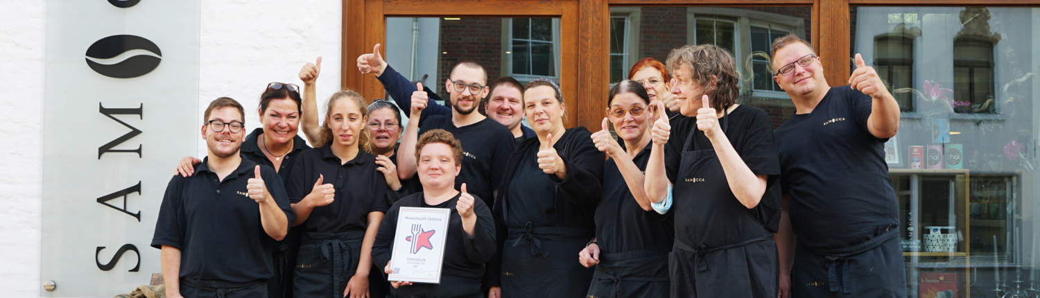 Gruppenfoto des Teams eines Restaurants vor dem Eingang, alle tragen schwarze Kleidung und zeigen Daumen hoch.